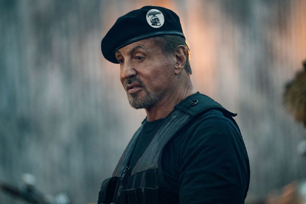 image | GotchaMovies Man wearing a black beret with a circular patch, looking to the left, in a dark vest against a blurred outdoor background.