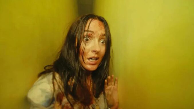 Woman with wide eyes and messy hair looks terrified in a yellow-lit corridor, hands raised defensively, face smeared with dirt or gore.