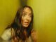 Woman with wide eyes and messy hair looks terrified in a yellow-lit corridor, hands raised defensively, face smeared with dirt or gore.