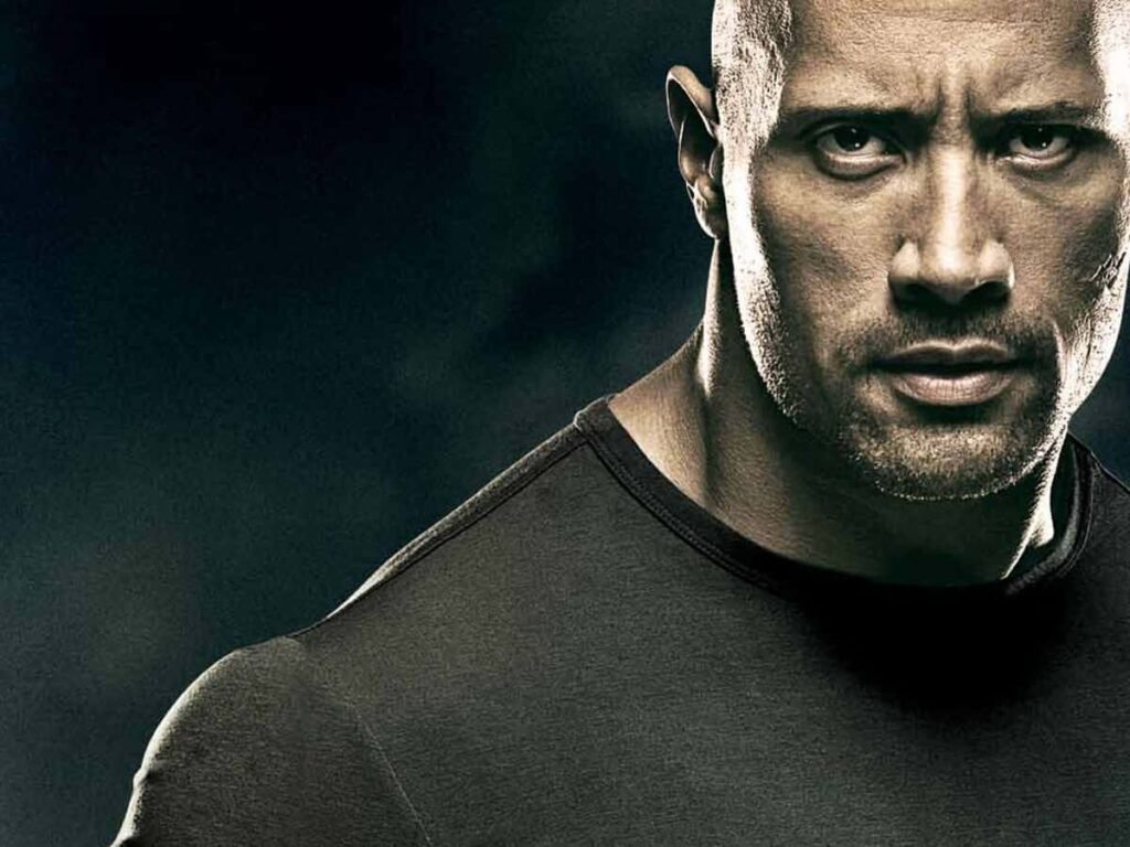 image | GotchaMovies Close-up of a muscular man with a stern expression wearing a dark athletic shirt against a dark background.