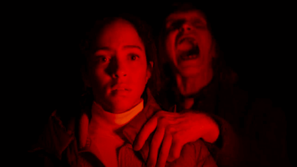 Two people in a dark scene lit by red light; the front person looks afraid while a second person behind screams and grips their shoulder.