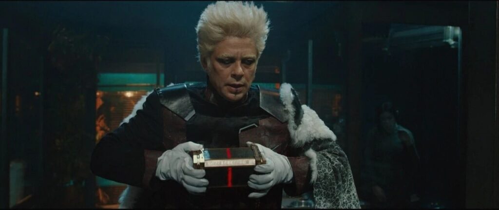 Person with spiky white hair in dark armor and a white fur cape, inspecting a small ornate box with a red stripe.