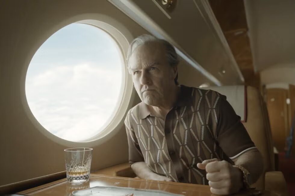 image | GotchaMovies Older man sits at a plane's dining table by a window, gazing forward with a glass of whiskey on the table.