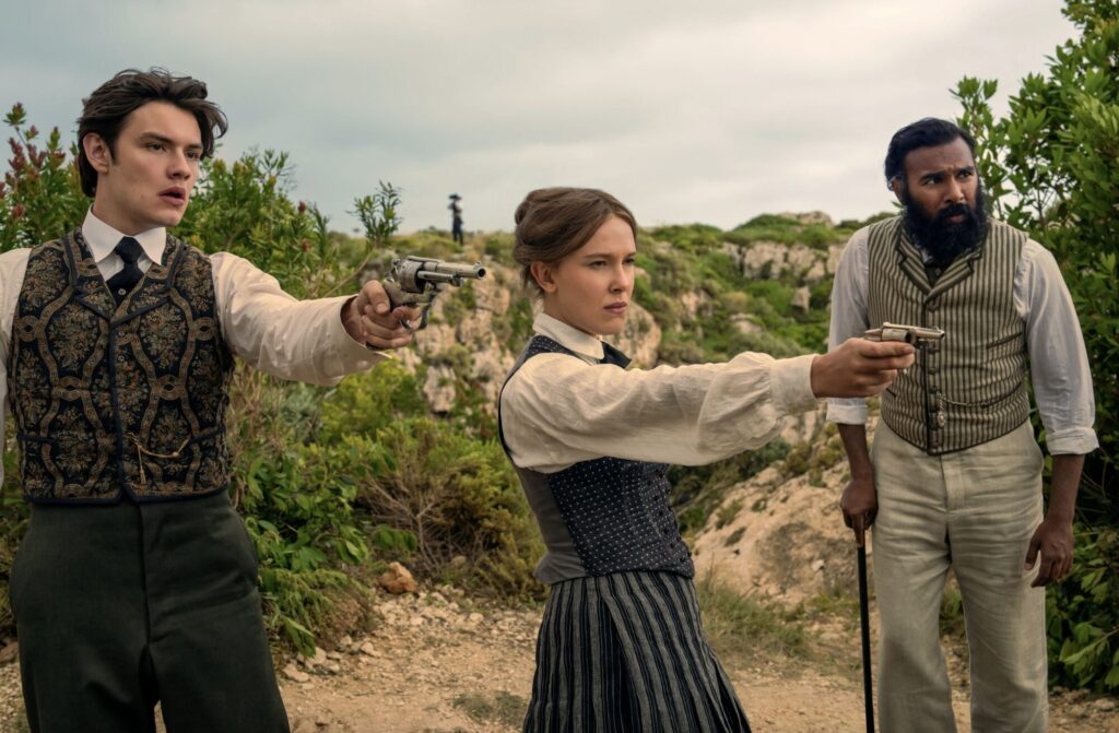 Three people in vintage clothing stand in a rocky outdoor setting, each holding a revolver and aiming off-frame, expressions tense.