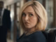 Blonde woman in a navy top sits in an office, looking to her left with a pursed lips expression.