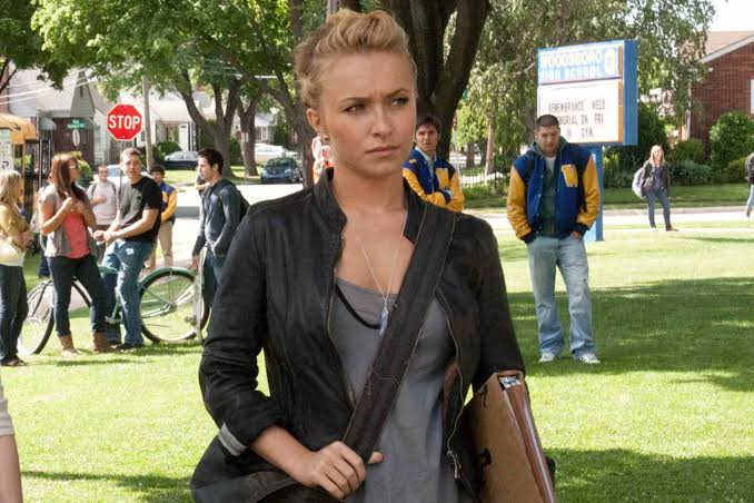 image | GotchaMovies Young woman with blonde hair in a black leather jacket and gray top, crossing a campus lawn with a brown messenger bag; students in the background.