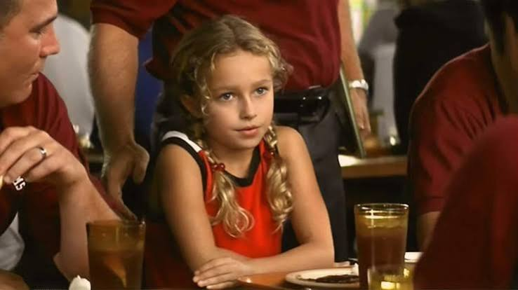 image | GotchaMovies Young girl with blonde braids wearing a red dress sits at a table with drinks, looking ahead thoughtfully.