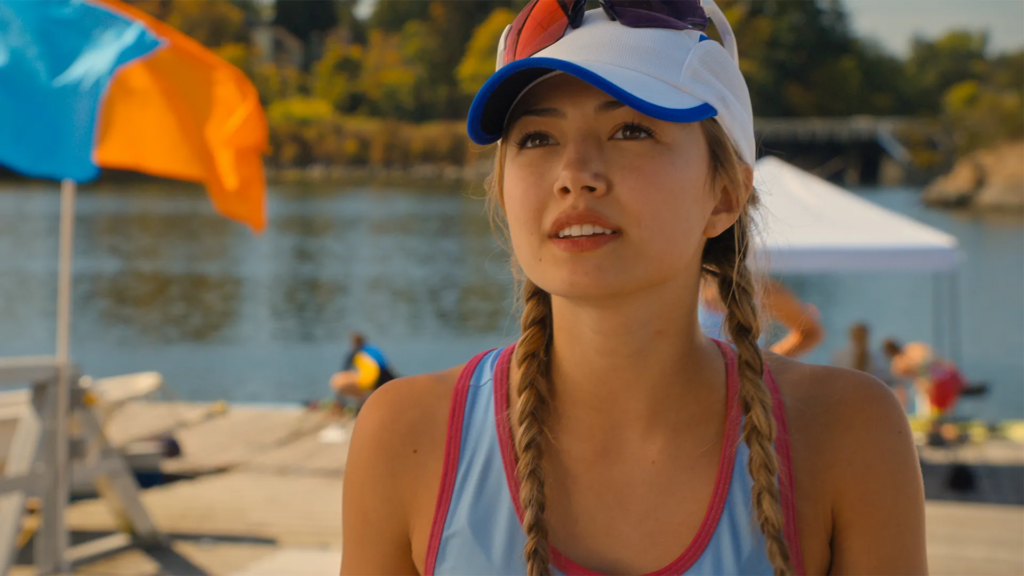 Young woman with braided pigtails, wearing a white-and-blue cap and pink-trimmed blue top, at a sunny lakeside beach.